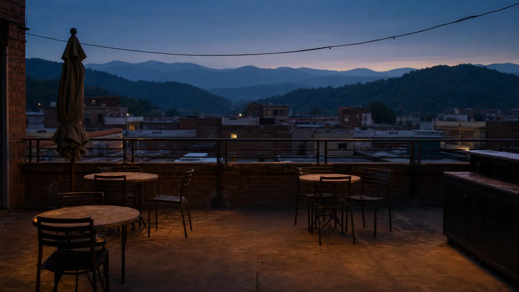 An empty rooftop terrace at blue hour above a low brick downtown, layered forested ridgelines receding into haze behind it.