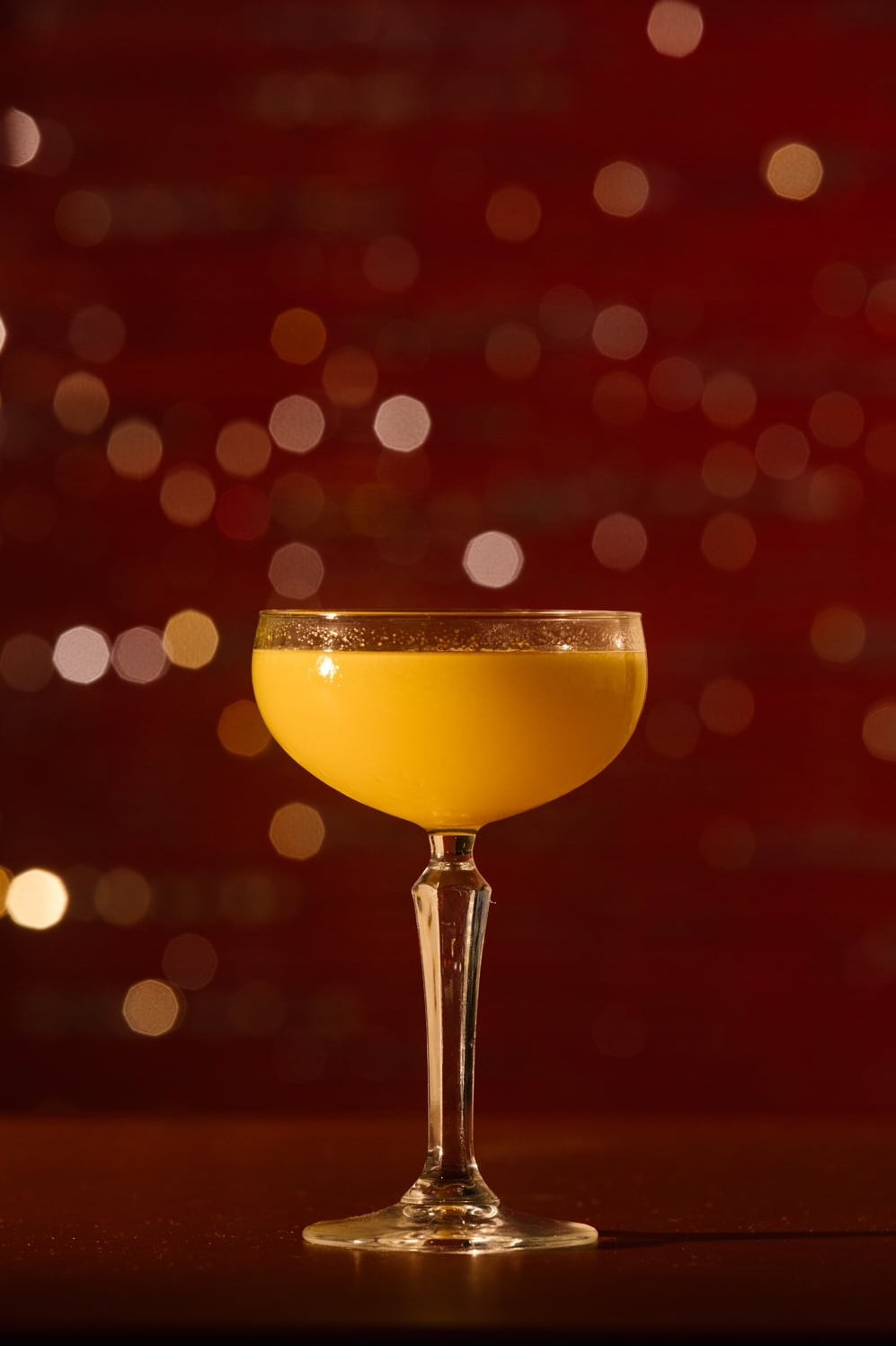 Golden cocktail in a coupe glass glowing against warm ambient bokeh lighting.