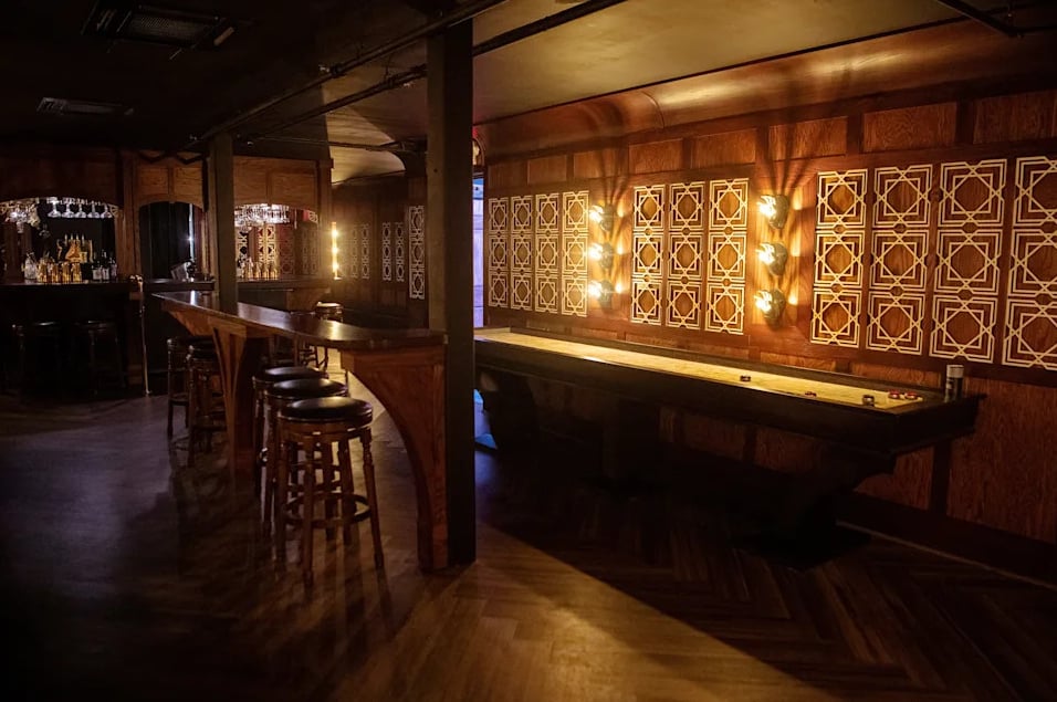Full room view with shuffleboard table, bar stools, and warm Art Deco lighting