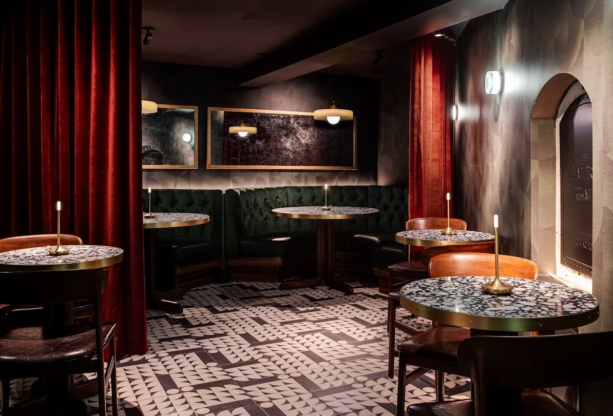 The intimate interior features plush green banquettes, rich red drapes, and a striking geometric tile floor.