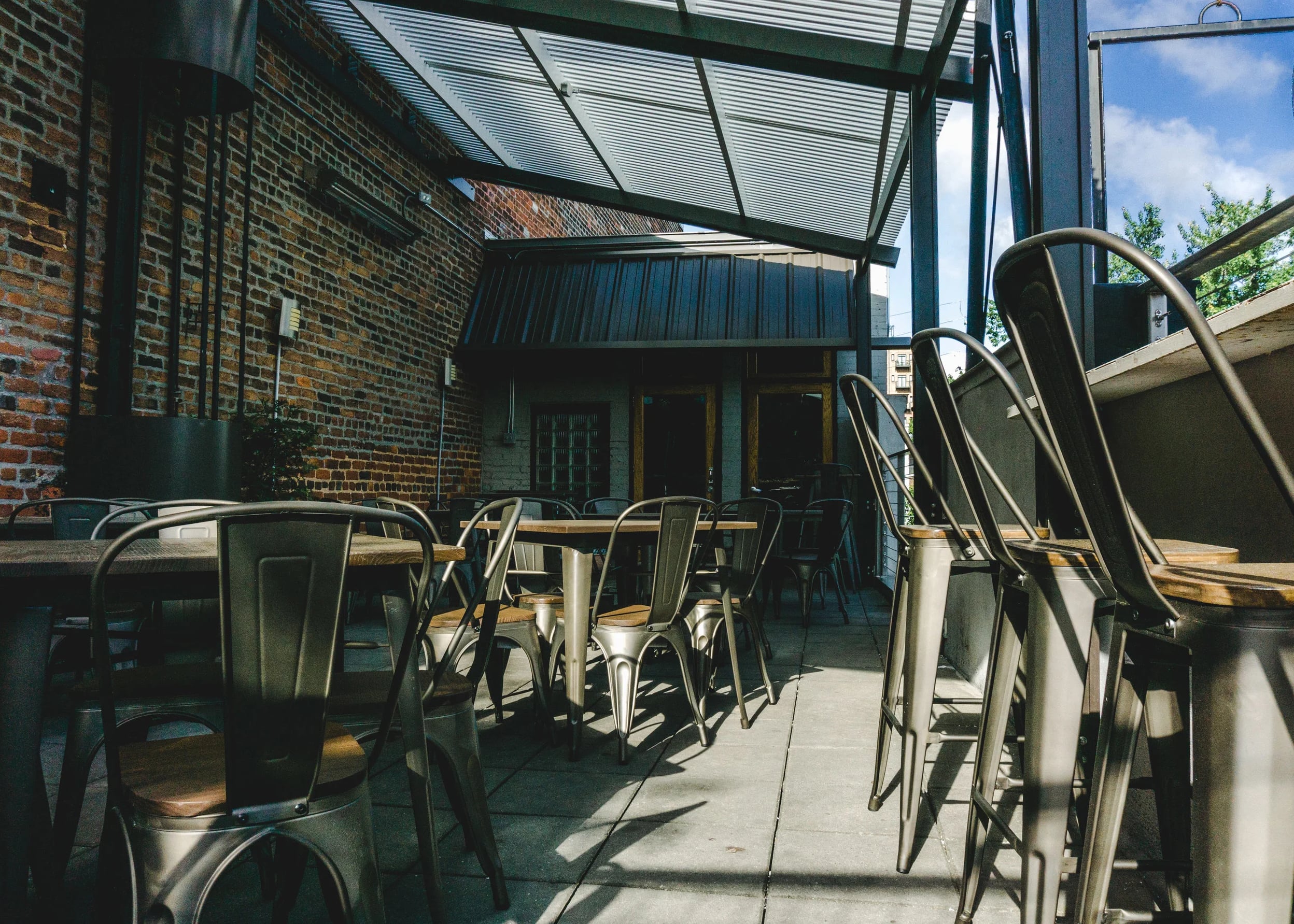 Industrial-style rooftop seating area with metal chairs and exposed brick wall under the louvered glass canopy.
