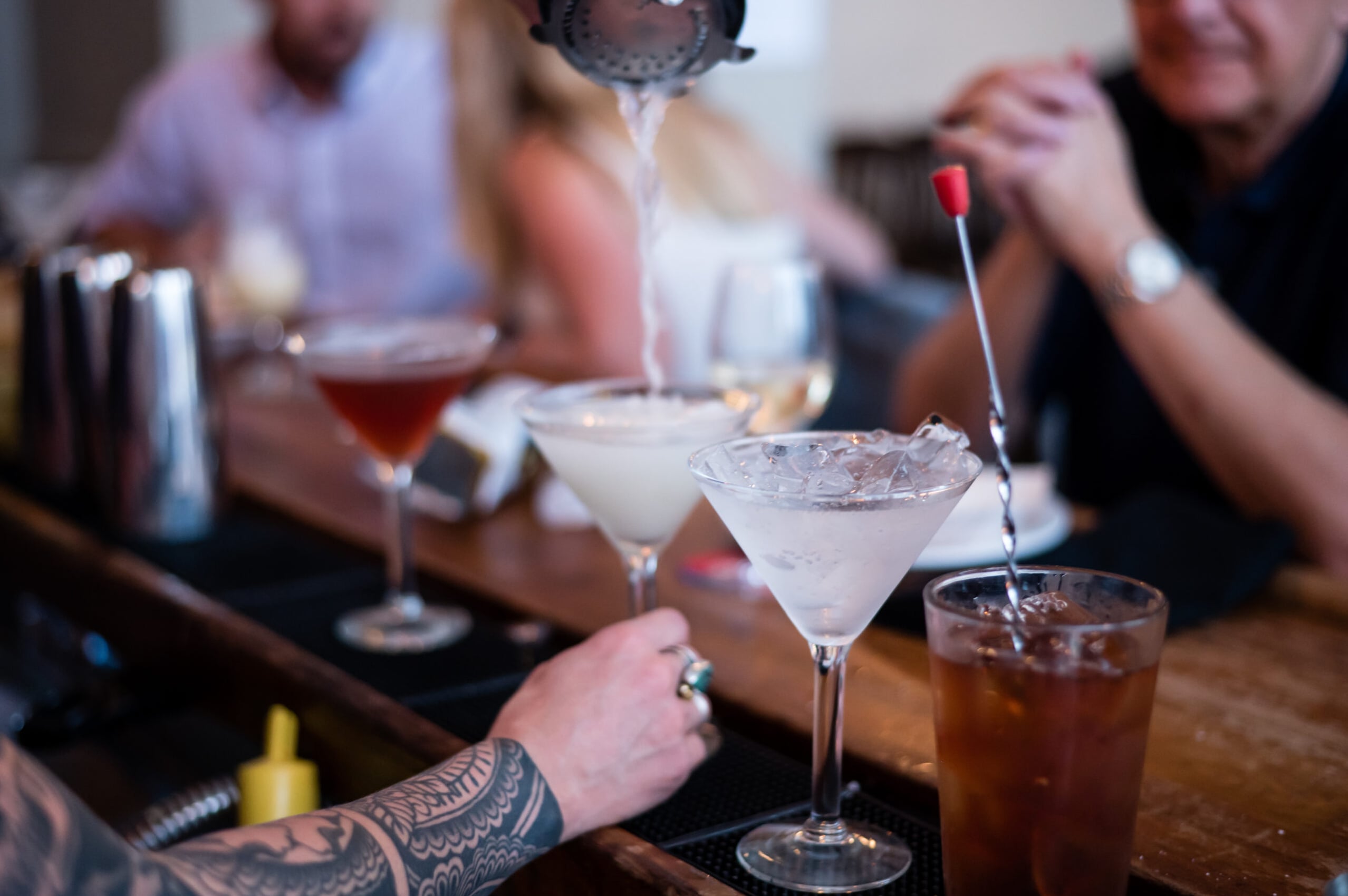 Bartender pouring a classic cocktail at the mahogany bar with patrons in the background.