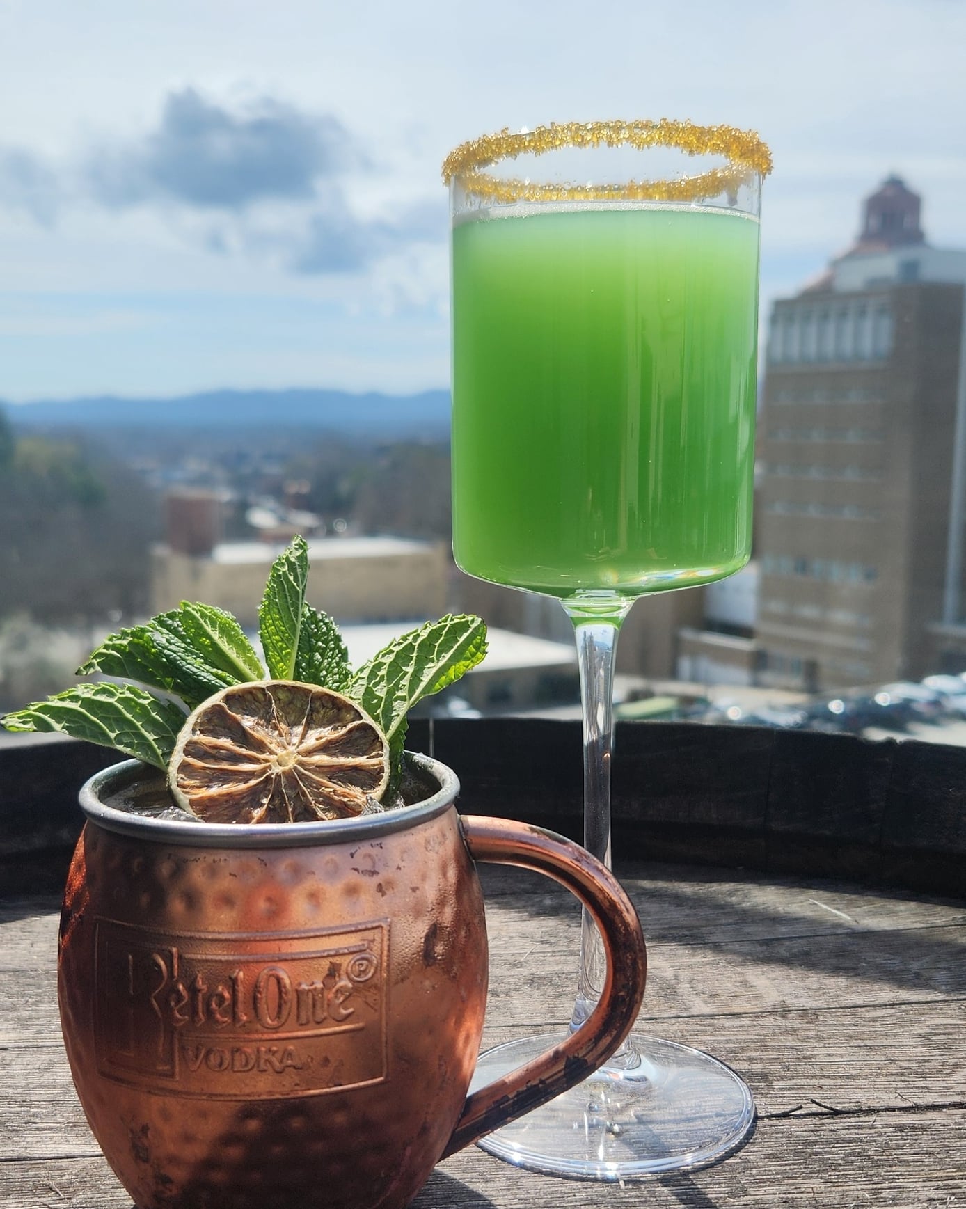 Signature green cocktail and Moscow Mule featuring a scenic city skyline view.