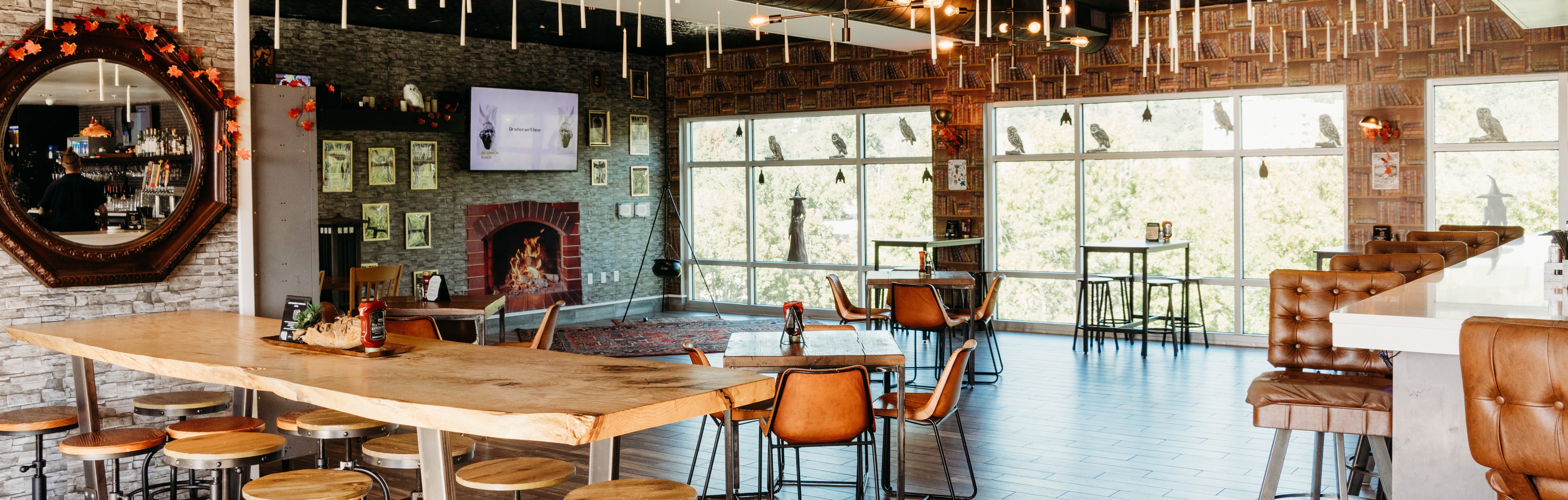 Bright industrial-chic indoor lounge with live-edge wood communal table, leather seating, exposed brick fireplace, and floor-to-ceiling windows
