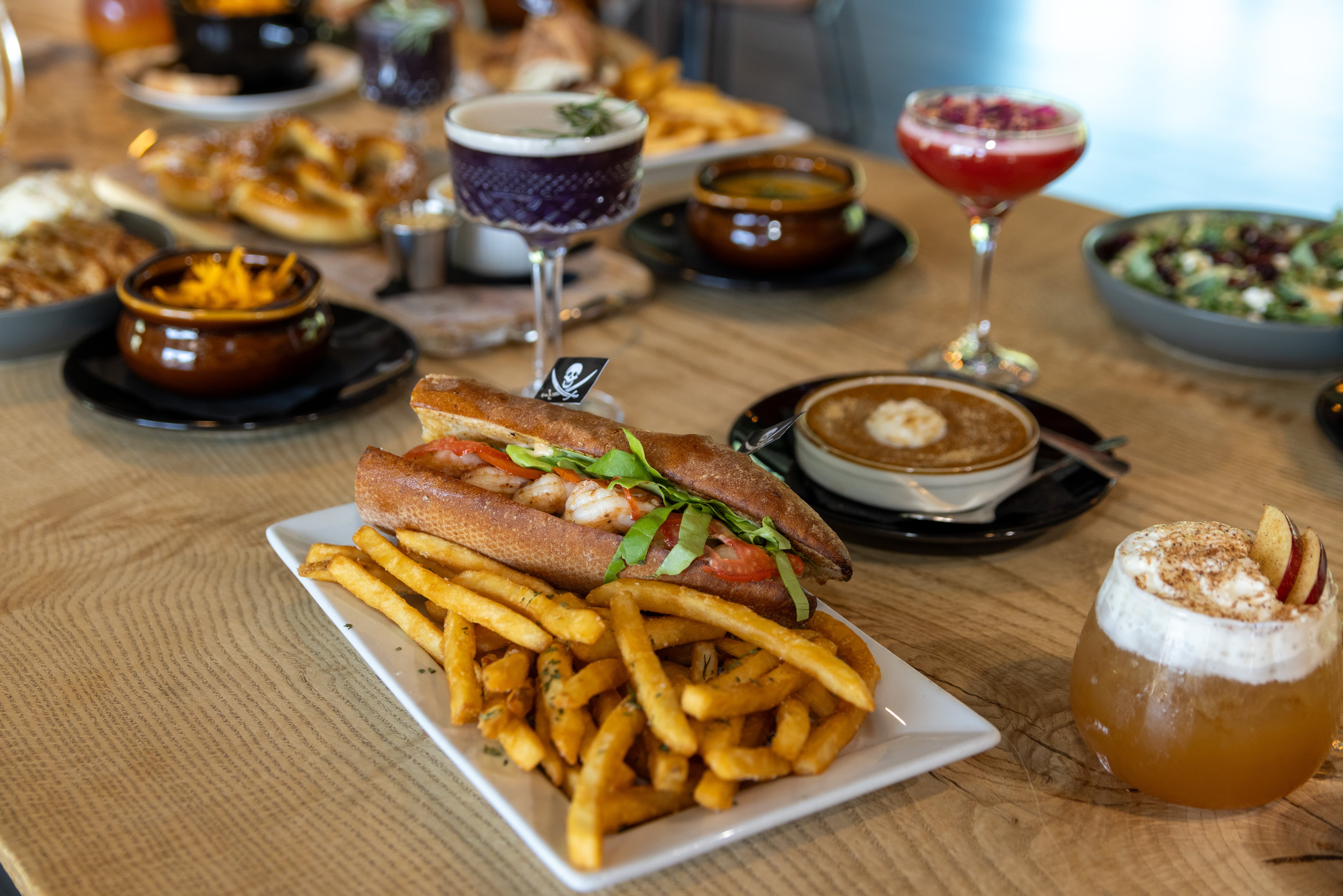 Pillar Kitchen spread — BLT sandwich with seasoned fries alongside cocktails including a blackberry coupe and apple mule