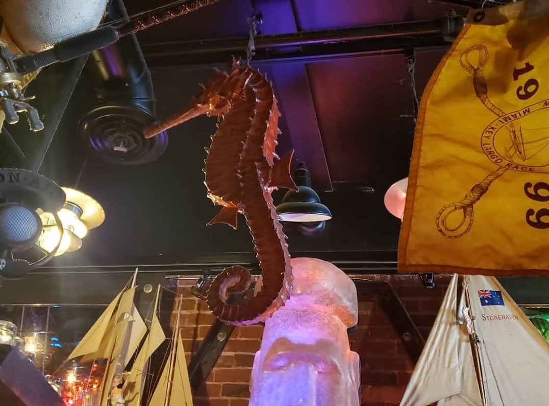 Large seahorse sculpture suspended from the ceiling amid nautical flags — part of the bar's eclectic maritime decor