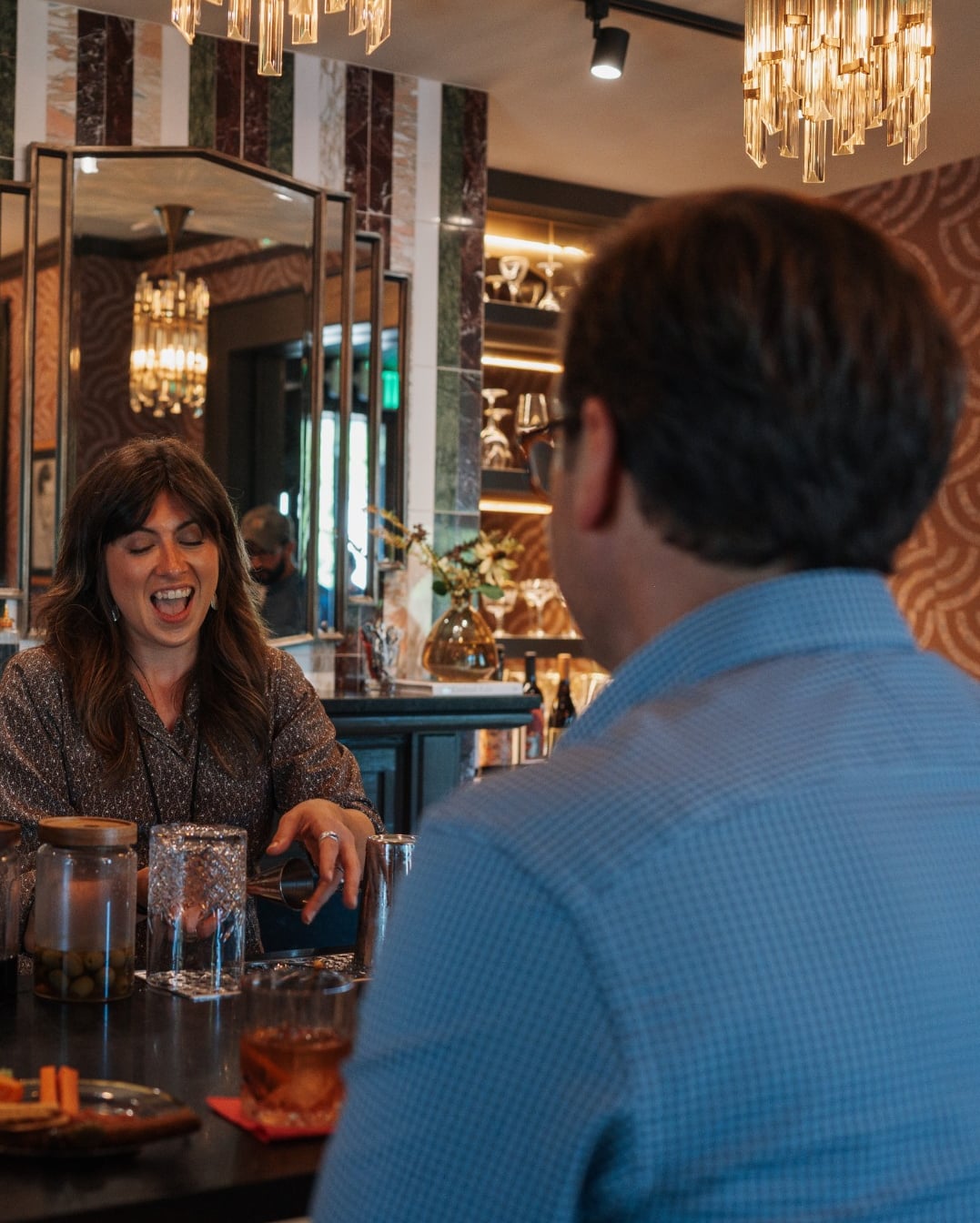 Bartender laughing and pouring drinks at an intimate cocktail bar with modern fixtures.