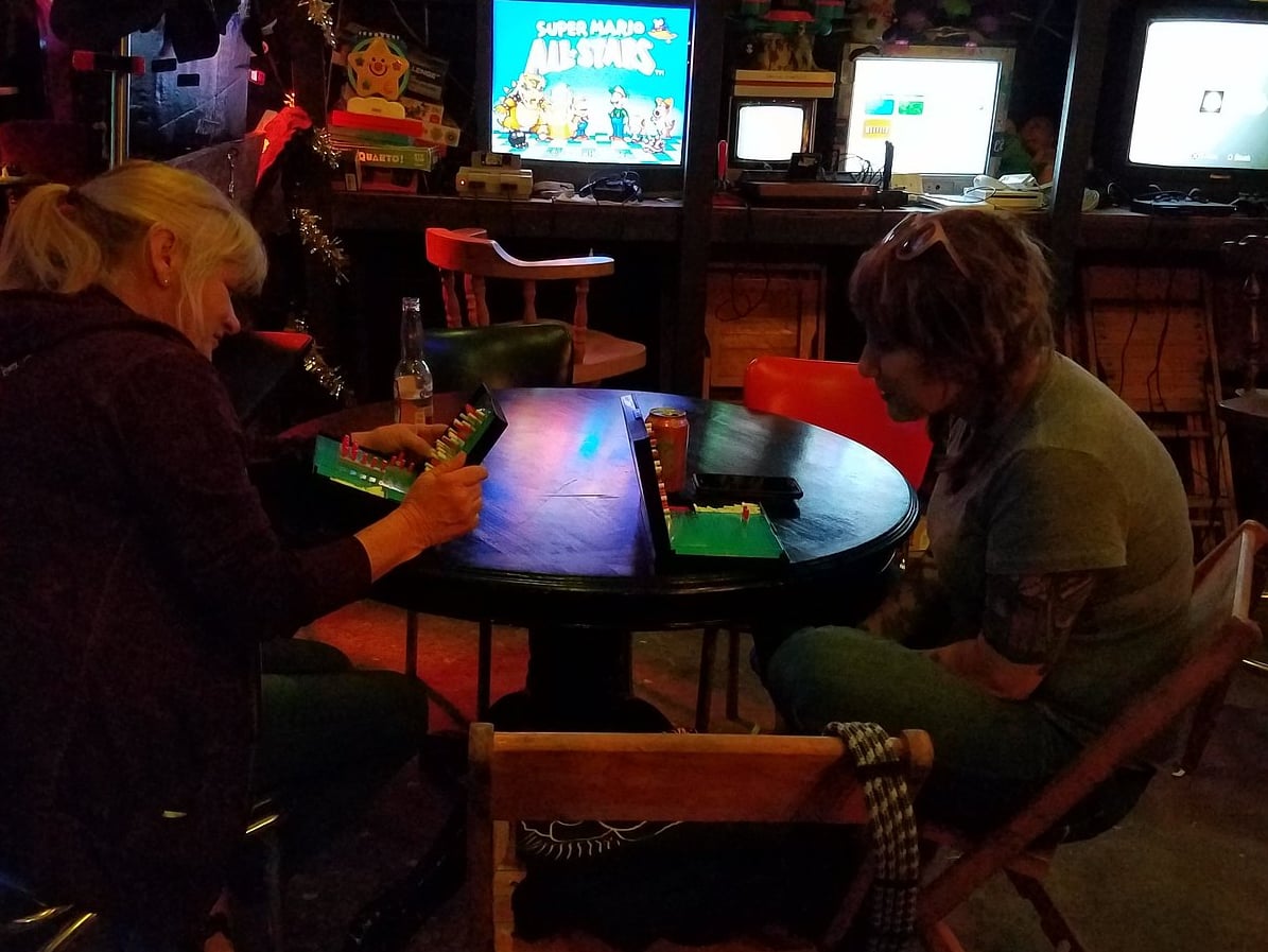 Guests playing board games at a vintage arcade table, surrounded by the lounge's wall of retro game consoles and TVs